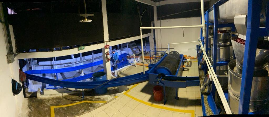 An industrial interior scene showing a large blue conveyor or processing line running through a warehouse-like space with tiled floors, white support columns, and metal machinery on the right side.